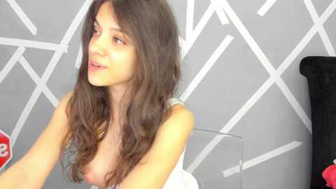 Media: Video of a smiling, light-skinned woman with long, wavy brown hair, wearing a white tank top, seated on a black chair, against a geometric, gray wall.