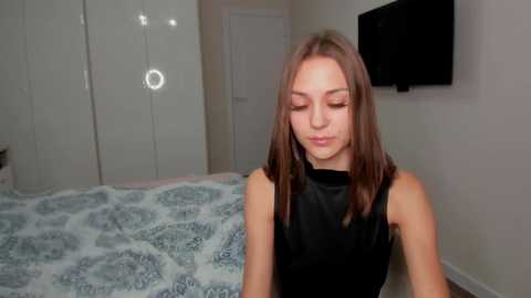 Media: Video of a young woman with light skin and straight, shoulder-length brown hair, wearing a black sleeveless top. She sits on a bed with a patterned duvet in a simple, beige-colored room with a flat-screen TV mounted on the wall.