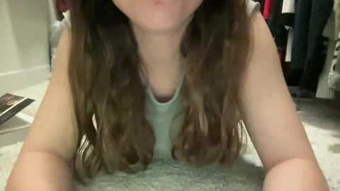 Media: Video of a light-skinned woman with long, wavy brown hair, wearing a light gray tank top, lying face-down on a carpeted floor in a cluttered, dimly lit room.