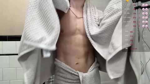 Media: Video of a shirtless, light-skinned male with a toned, muscular abdomen, wearing a white towel around his waist. Background includes a tiled bathroom wall with white tiles and a black border.