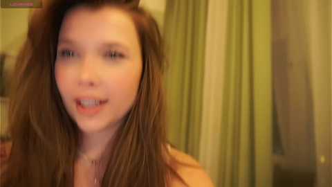 Media: A blurry video of a young woman with long, straight brown hair, wearing a light-colored top, standing indoors with beige curtains and green walls in the background.