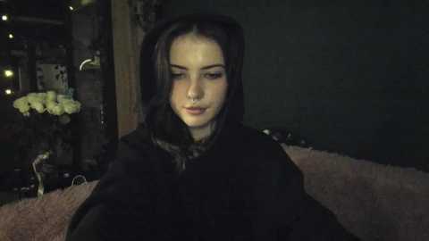 Media: Video of a young woman with pale skin, long dark hair, and a black hooded cloak, sitting on a plush pink sofa in a dimly lit room with a wooden background and a white floral arrangement.