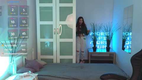 Media: Video of a young woman with long, curly hair in a green shirt and ripped jeans, standing in a modern, blue-themed bedroom with a large bed, white wardrobe, and a digital clock displaying 4:50.