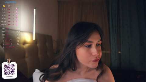 Media: A video of a Latina woman with long dark hair, wearing a white strapless dress, gazing contemplatively. Background includes a dimly lit room with a large bed and window curtains.