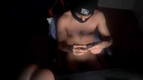 Media: Video of a shirtless man with a black cap, focused on a smartphone, in dim lighting, sitting on a black chair, with a blurred background.