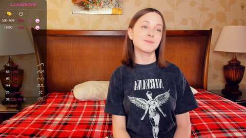 Media: Video of a young woman with straight brown hair, light skin, and a slim build, wearing a black Metallica t-shirt. She sits on a red plaid bedspread in a cozy bedroom with wooden headboard, vintage lamps, and floral wallpaper.