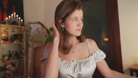 Media: Video of a young woman with light skin and shoulder-length brown hair, wearing a white off-shoulder dress, standing indoors, looking thoughtful. Background includes a bookshelf and lit candles.