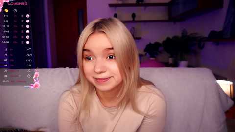 Media: Video of a young, fair-skinned, blonde woman with a slight smile, wearing a beige sweater, seated on a white couch in a dimly lit living room.