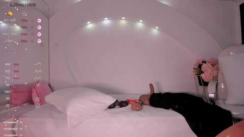 Media: A video of a man in black clothes lying on a white bed in a white, minimalist bedroom, with a pink vase of flowers on the nightstand.