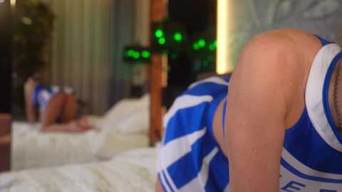 Media: Video of a woman in a blue and white striped sports bra and shorts, bending over a bed, blurred background with green lights.