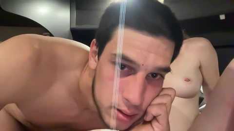 Media: A video of a shirtless man with short black hair, light skin, and a slight beard, lying on his side, looking at the camera. In the background, a partially visible nude woman with fair skin and small breasts.