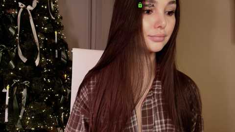 A video of a young woman with long, straight, dark brown hair, wearing a brown plaid shirt, standing beside a decorated Christmas tree with white ribbons.