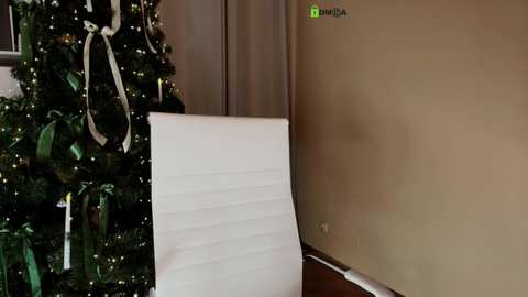 Media: Video of a beige wall with a white, ribbed office chair, partially visible in the corner, next to a decorated Christmas tree with green ornaments, lights, and a white ribbon.