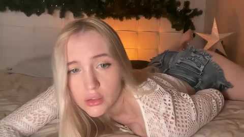 Media: Video of a fair-skinned, blonde woman with a pouty expression, wearing a white crochet top and denim shorts, lying on a bed in a cozy, festive room with a lit Christmas tree and garland in the background.