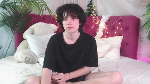 Media: Video of a young, androgynous person with short black hair, pale skin, and a neutral expression, wearing a black T-shirt, sitting on a bed with white sheets and a pink headboard.