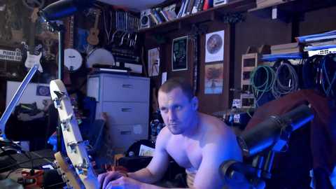 Media: Video of a shirtless, muscular man with short hair, sitting at a cluttered desk in a dimly lit, chaotic room filled with musical instruments, cables, and posters.