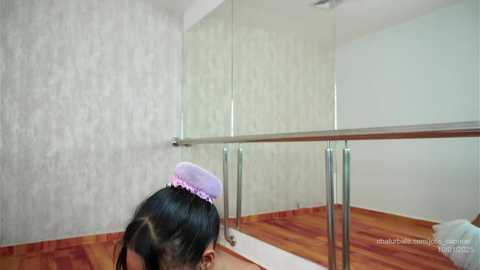 Media: Video of a young girl with dark hair wearing a pink headband, lying on a hardwood floor in a room with light gray textured wallpaper and a mirrored ballet barre.