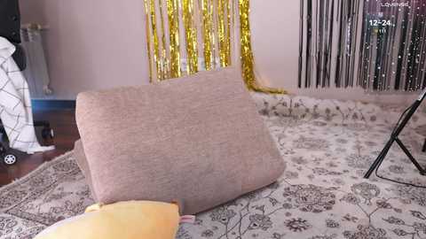 Media: A video of a cozy living room featuring a beige cushion on a floral-patterned rug, with gold tinsel and a silver star backdrop, and a black tripod camera on the right.