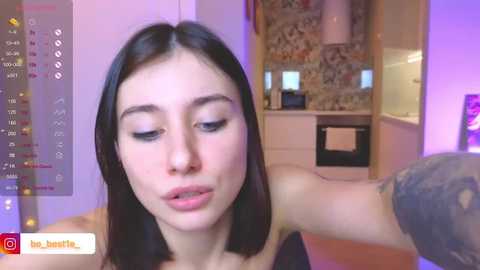 Media: Video of a young woman with straight black hair and fair skin, wearing a black top, in a modern kitchen with floral wallpaper and purple lighting.