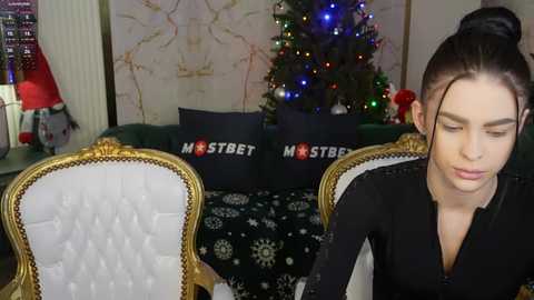 Media: Video of a young woman with dark hair in a bun, wearing a black dress, sitting in a vintage-style chair with gold trim, in a cozy room with a decorated Christmas tree and \"Mister Street\" pillows.