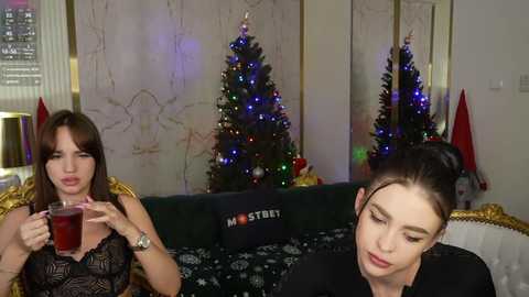 Media: Video of two women, one with long brown hair, wearing a black lace top, holding a glass; the other with dark hair, wearing a black top, sitting on a dark couch with a festive background.