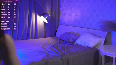 Media: A video of a dimly lit bedroom with a white bed, gray sheets, and a purple light illuminating a wall-mounted lamp.