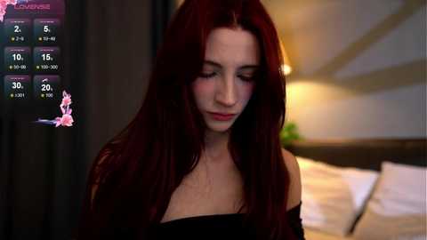 Media: Video of a young woman with long, straight, deep red hair, wearing a black off-shoulder top, standing in a dimly lit bedroom with a bed in the background.