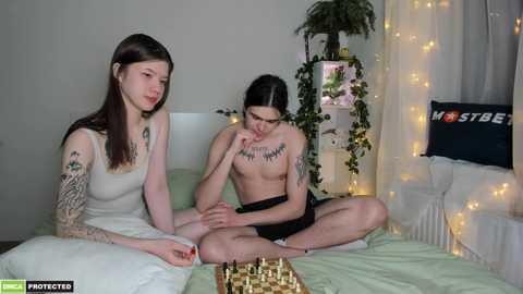 Media: Video of two young women sitting on a bed in a cozy, dimly-lit room. One wears a white tank top, the other is topless with a tattoo on her chest.