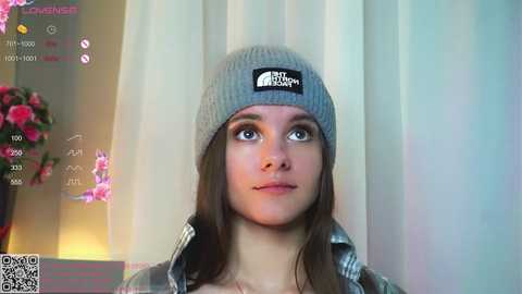 Media: Video of a young Caucasian woman with long brown hair, wearing a grey beanie with a black logo, a plaid shirt, and a neutral expression, against a background of pink flowers and white curtains.