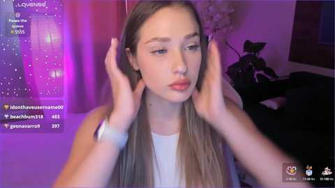 Media: Video of a young Caucasian woman with light skin and long, straight blonde hair, wearing a white tank top, touching her hair with both hands. Background features a purple and pink gradient, a potted plant, and a camera.