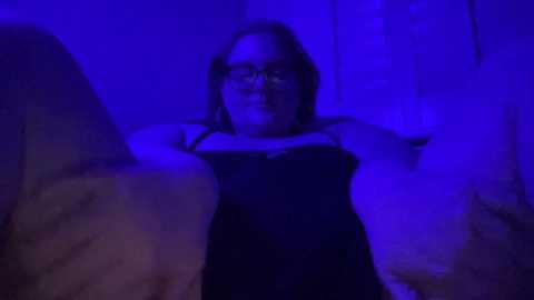 Media: A dimly lit video of a woman with glasses, fair skin, and shoulder-length hair, wearing a black lace bra and a black dress, standing with arms raised, bathed in blue light.