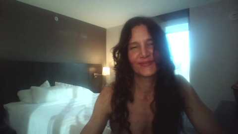 Media: Video of a smiling woman with long, wavy brown hair, wearing a black lace top, standing in a dimly lit, modern bedroom with a neatly made bed and a window with white curtains.