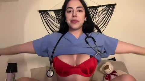 Media: A video of a young woman with long dark hair, fair skin, and a red bra, wearing blue scrubs and a stethoscope, sitting on a beige bed with a black hammock and teddy bear in the background.