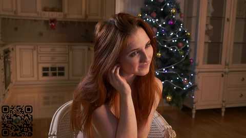 Media: Video of a fair-skinned woman with long, wavy auburn hair, sitting indoors, wearing a white top, smiling gently while looking off-camera. Christmas tree with colorful lights and ornaments in the background, wooden cabinets, and a QR code on the left.