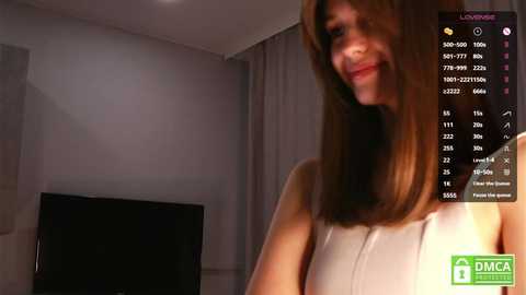 Media: A video of a smiling, light-skinned woman with shoulder-length brown hair, wearing a sleeveless white top, standing in a modern, dimly lit room with a TV and gray curtains.