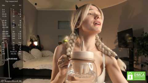 Media: Video of a fair-skinned woman with blonde braided pigtails, wearing a white tank top, holding a glass jar, in a cozy bedroom with a bed, TV, and clock.