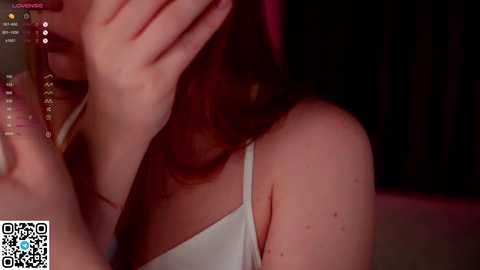 Media: Video of a woman with long, dark hair, wearing a white tank top, covering her face with one hand, in a dimly lit room.