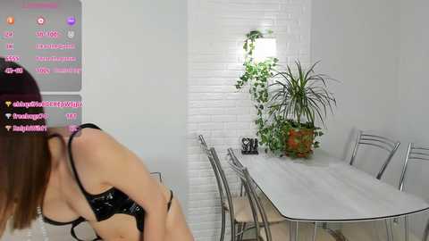 Media: A video shows a woman in a black bra, leaning over a table, in a modern, white-walled room with a white brick wall and potted plants.