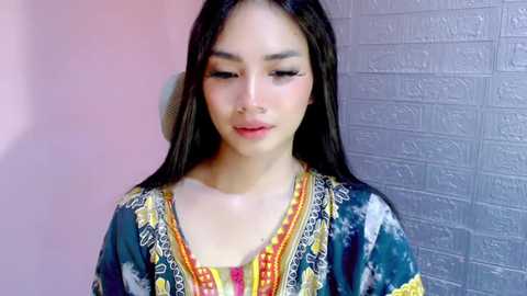 Media: Video of a young East Asian woman with long black hair, fair skin, and delicate features, wearing a colorful, patterned top against a textured, gray wall background.