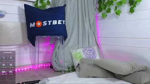 Media: A video of a cozy bedroom with white wooden walls, a blue pillow with \"MOSTBET\" text, a green blanket, a gray cushion, and purple mood lighting.