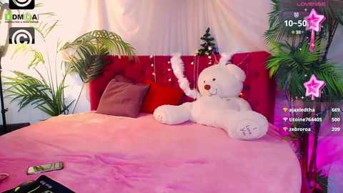 Media: Video of a plush, white teddy bear on a pink bedspread with red cushions, surrounded by green plants and a Christmas tree, with social media icons overlaying the image.