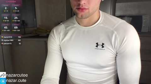 Media: Video of a fit, fair-skinned man with a short beard, wearing a tight, white Under Armour long-sleeve shirt. Background shows a dimly lit room with a TV screen displaying fitness stats.