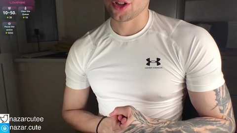 Media: Video of a muscular, tattooed man in a white Under Armour shirt, captured from the chest up, indoors.