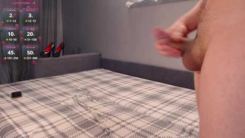 A video of a nude man with a large erect penis standing beside a bed with a plaid blanket and red high heels on the floor.