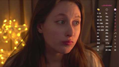 Media: Video of a young, fair-skinned woman with long brown hair, wearing a white top, looking contemplative. Background features warm, ambient lighting and a string of fairy lights.