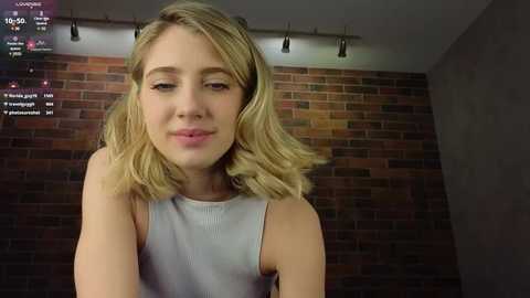 Media: Video of a young Caucasian woman with shoulder-length blonde hair, wearing a sleeveless gray top, against a brick wall background. Streaming video with chat comments visible in the top left corner.