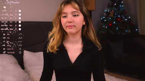 Media: A video of a young woman with shoulder-length blonde hair, wearing a black shirt, standing in a softly lit bedroom with a decorated Christmas tree in the background.