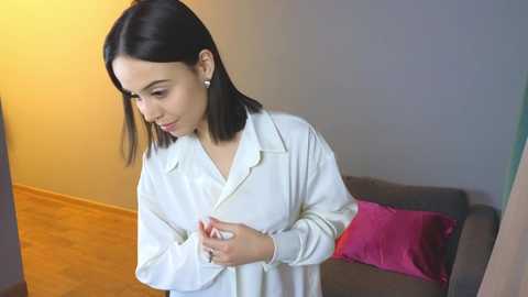 Media: Video of a young woman with straight black hair, wearing a white shirt, adjusting a necklace in a room with a beige wall and a pink pillow on a gray couch.