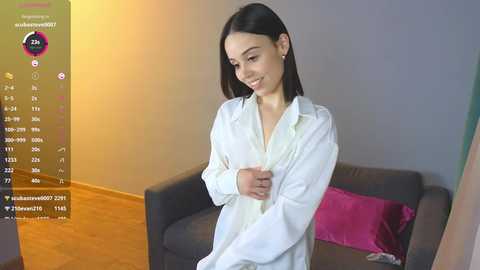 Media: Video of a smiling, slender young woman with medium-length dark hair, wearing a white button-down shirt, standing in a modern living room with a gray couch and pink pillow.