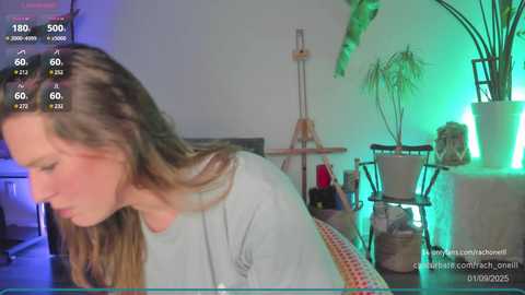 Media: Video of a young woman with long brown hair, wearing a light gray top, in a dimly lit, modern living room with green lighting, potted plants, and a piano.